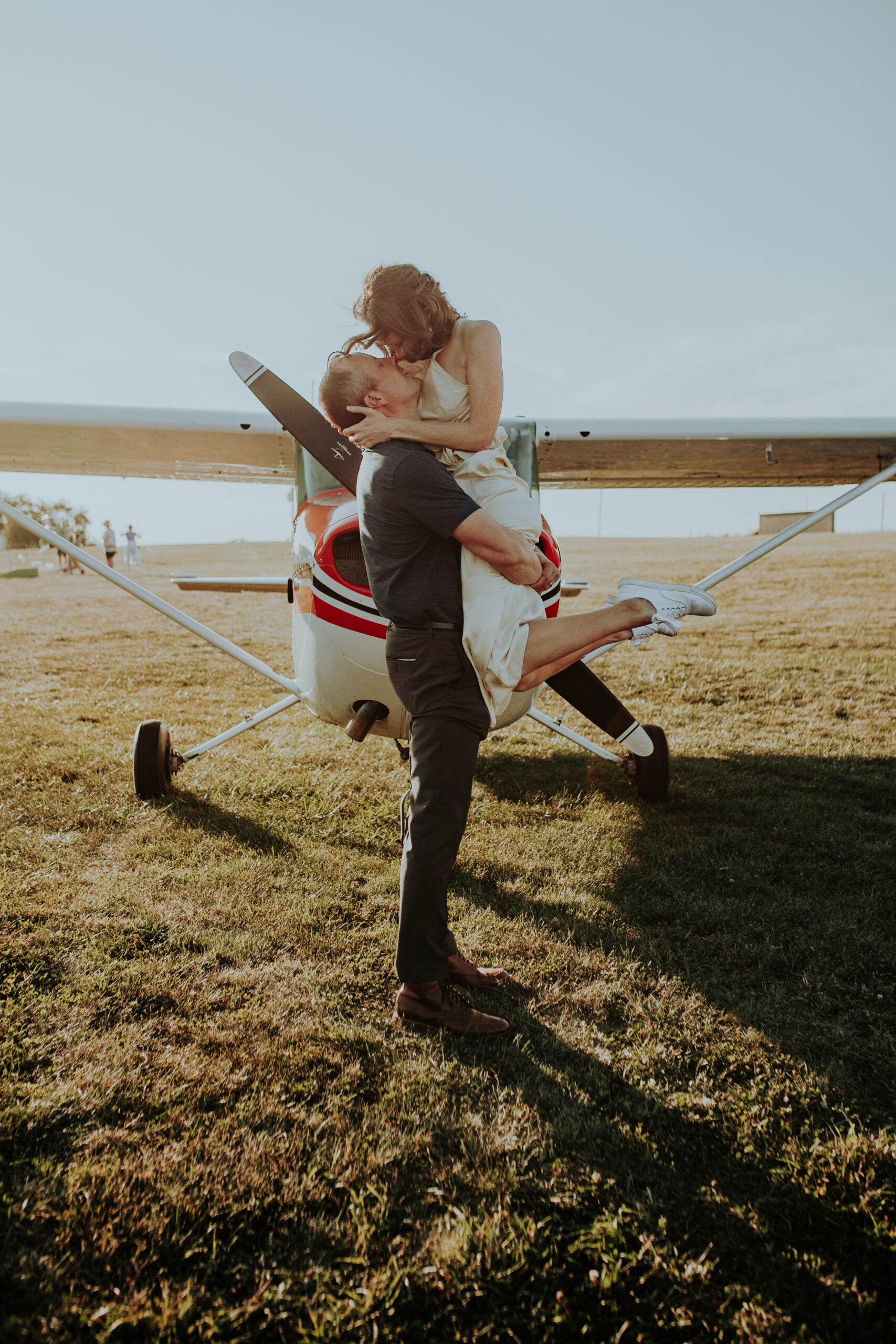 A groom lifts his bride off the ground in front of a small red and white airplane at Galt Airport. They share a kiss under the bright sun, with the bride wearing a simple dress and sneakers