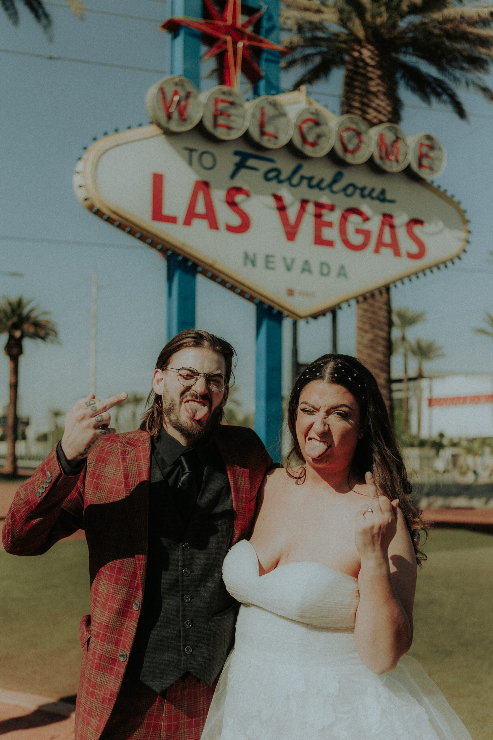 newlywed couple making playful faces and flipping off the camera in front of the iconic 'Welcome to Fabulous Las Vegas' sign. The groom wears a red plaid suit and the bride wears a white gown.