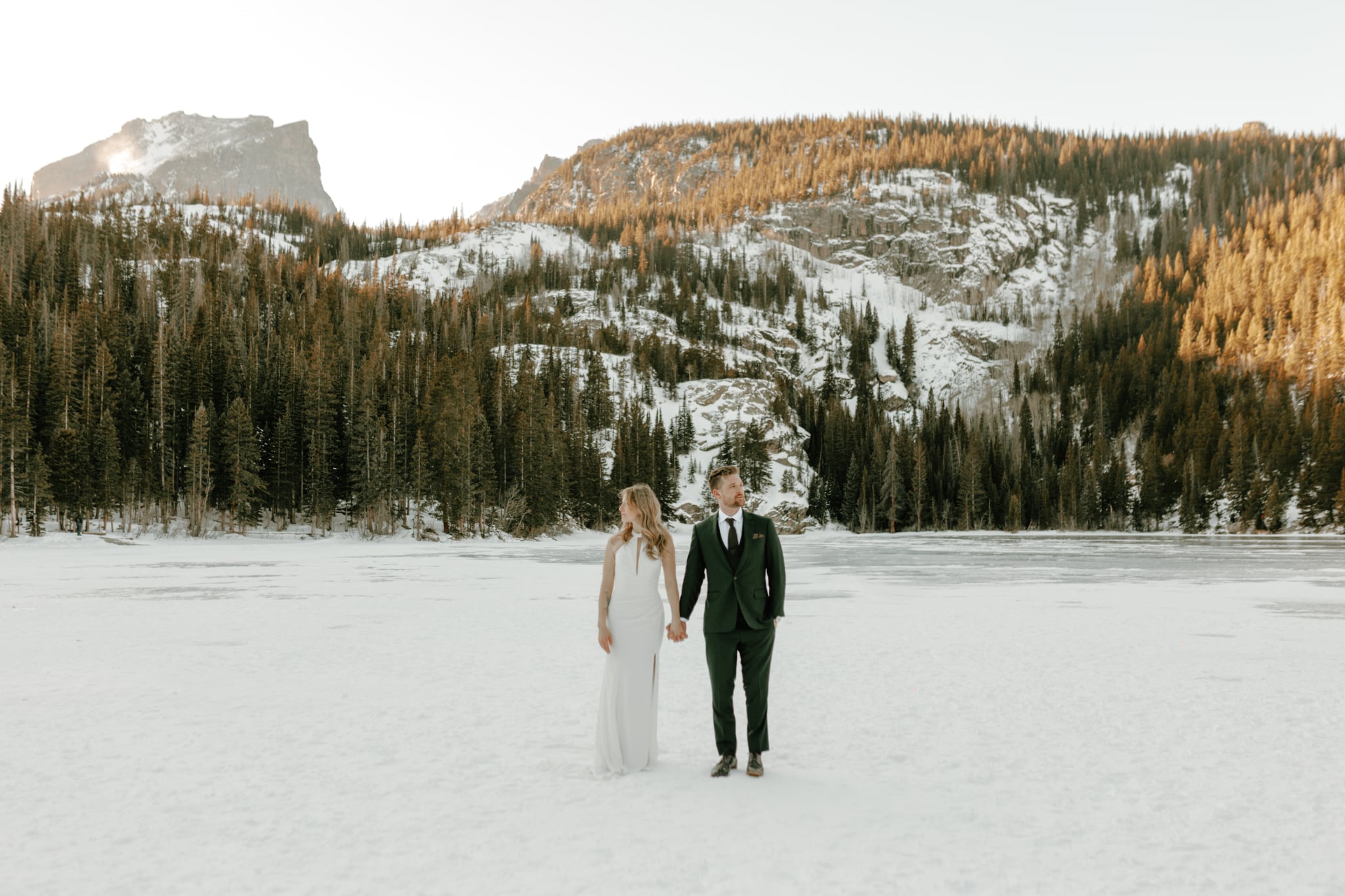 bride and groom holding hands walking across a frozen Bear Lake in Rocky Mountain National Park in winter with snow covered mountains and golden lit pine trees in the background