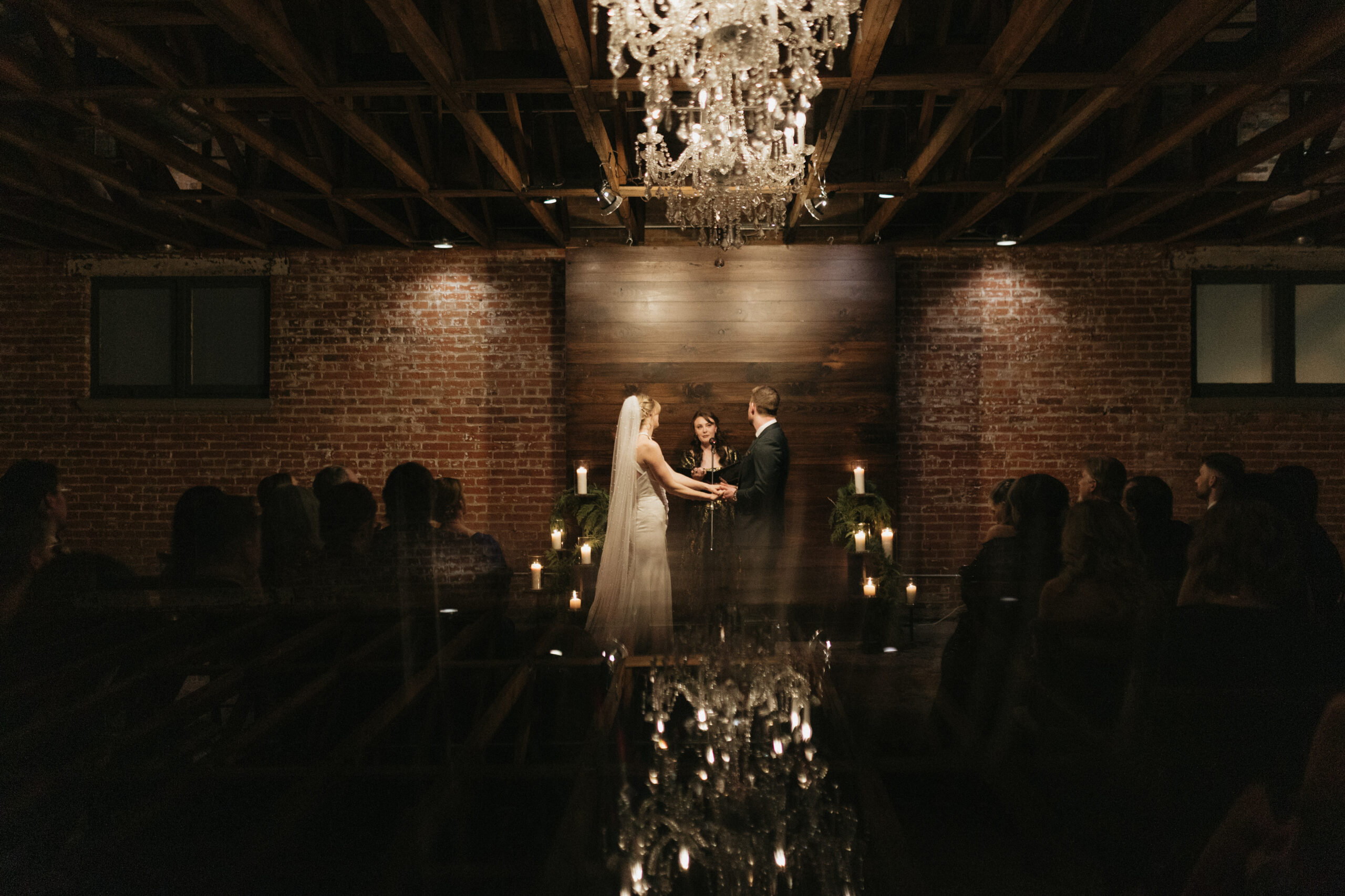ride and groom exchanging vows by candlelight at the St. Vrain in Longmont, Colorado, beneath a crystal chandelier with exposed brick walls and intimate guest seating on New Year's Eve.