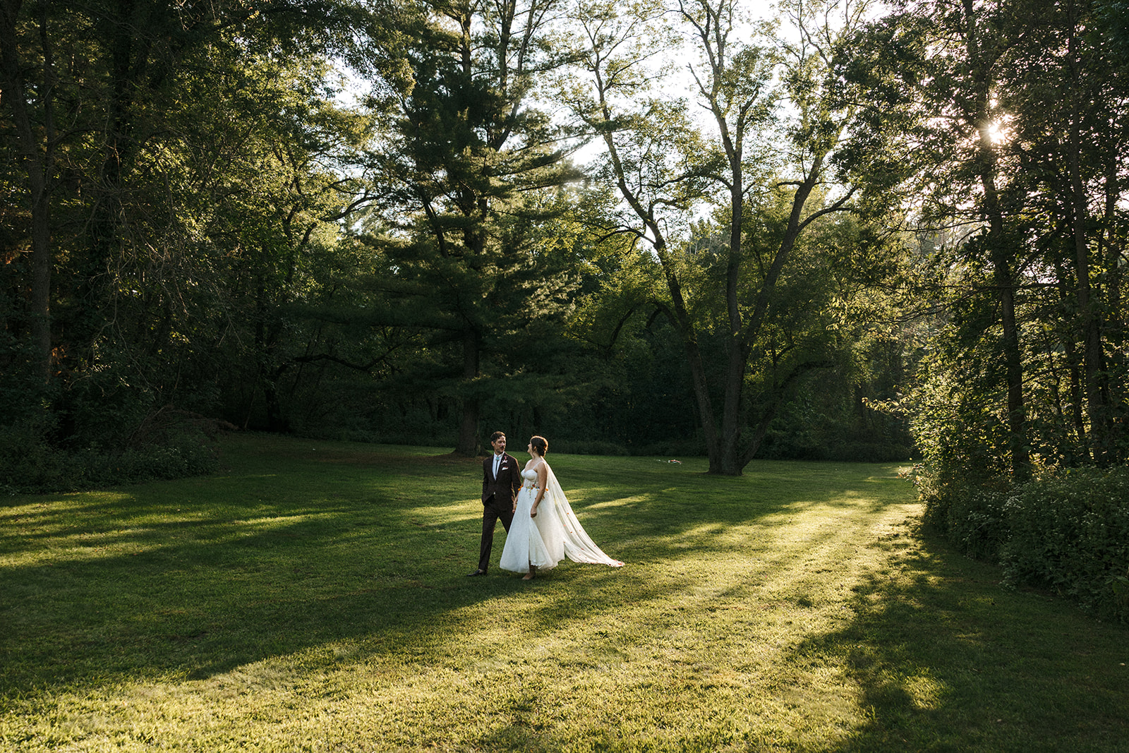 husband and wife walk hand in hand during sunset in open grass area surrounded by trees